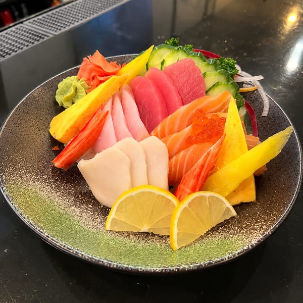 A bowl of assorted sashimi including tuna, salmon, yellowtail, and crab with cucumber, lemon, and pickled ginger on a green speckled plate.