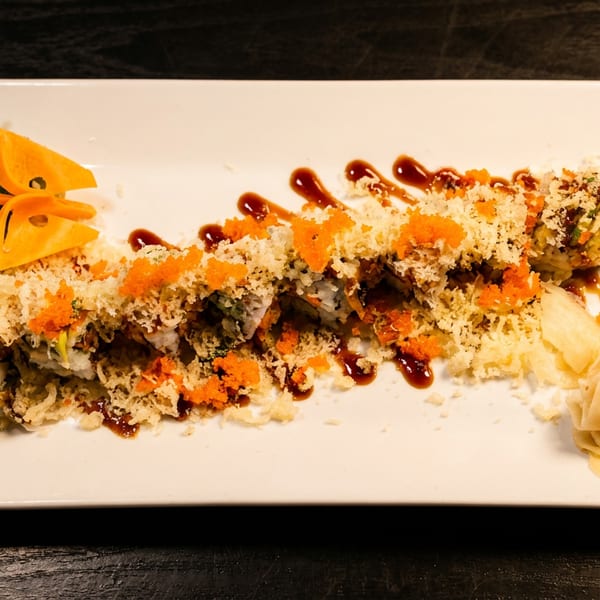 A long sushi roll topped with orange roe, tempura flakes, and eel sauce, garnished with pickled ginger and wasabi.